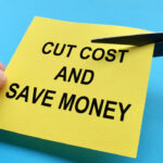 Scissors cutting yellow notepad with cut cost and save money text on blue background - Business Cost Saving Ideas