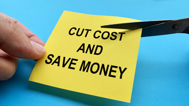 Scissors cutting yellow notepad with cut cost and save money text on blue background - Business Cost Saving Ideas